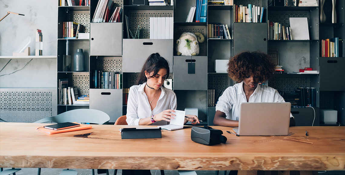 Two young women working together at a large home office desk, featuring a bright, spacious environment.