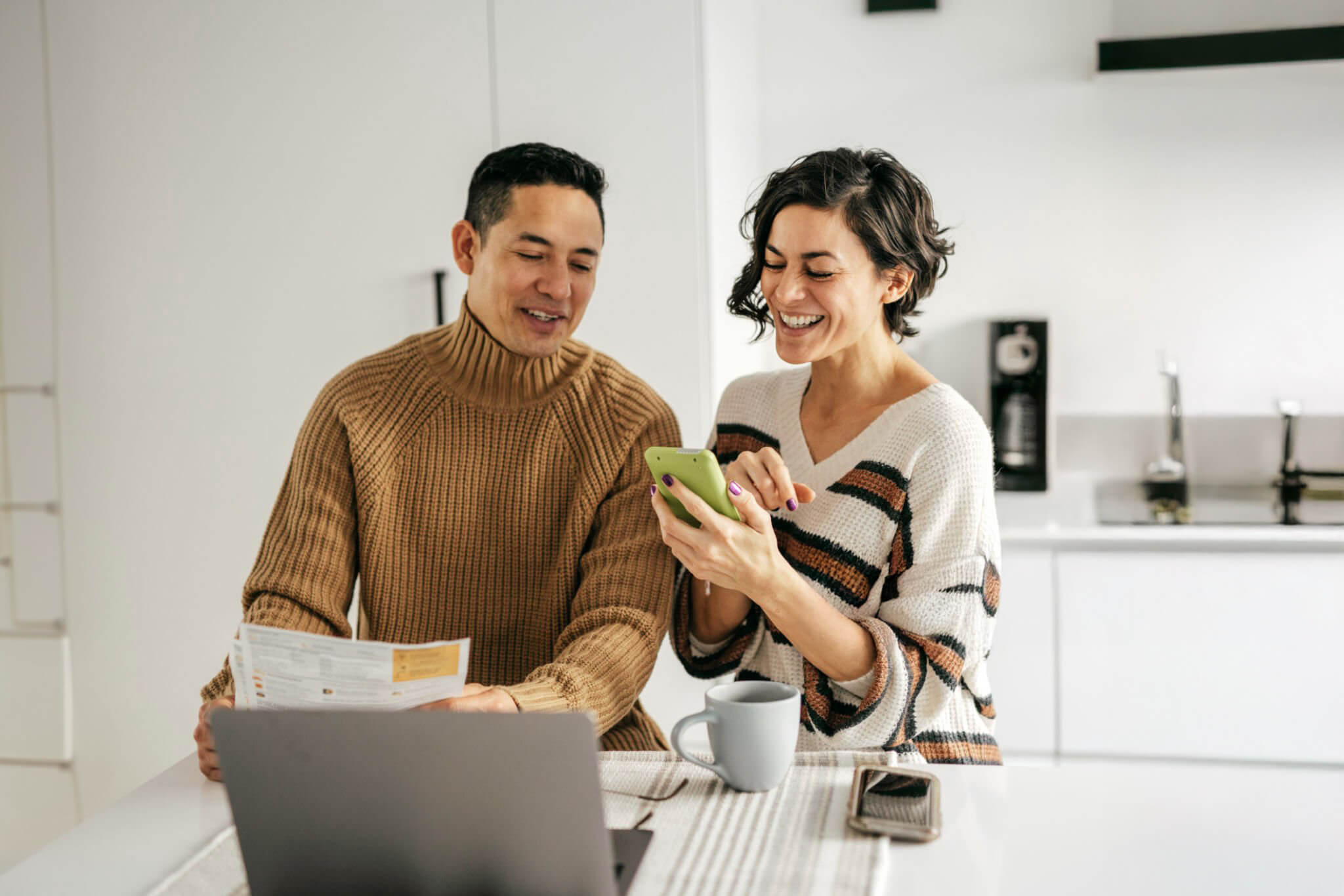 Smiling couple in their kitchen reviewing paperwork and a phone together while planning their first home purchase.